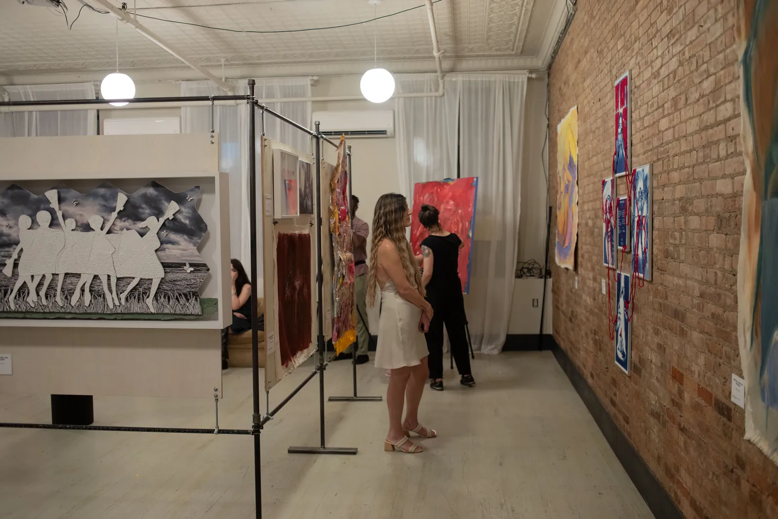 A visitor in a cream slip dress stands before artworks hung on an exposed brick wall, beside freestanding panels holding a large paper-cut work of white figures against a stormy sky.